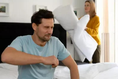 Mann sitzt auf Bett, Frau schwingt Kissen im Hintergrund, Schlafzimmer, Alltagsszene