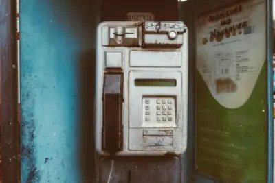 Ein alter, rostiger Telefonautomat in blauem Gehäuse mit einer grünen Werbetafel rechts.